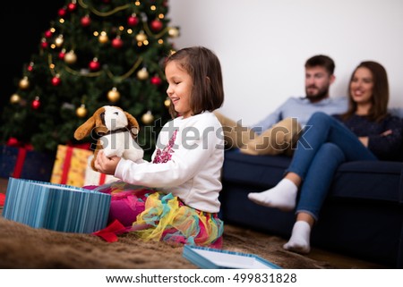 Happy little girl playing with her Christmas present stuffed puppy toy while sitting at home with her parents in front of the Christmas tree. Soft focus