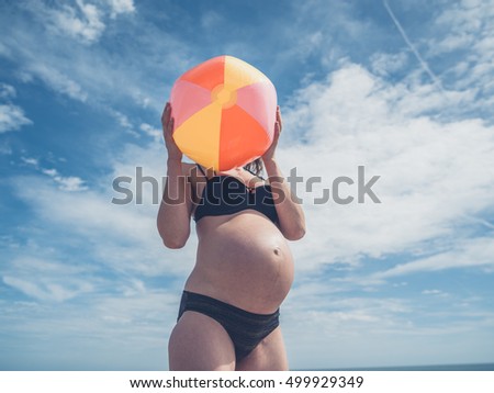 A young pregnant woman is standing outside with a beach ball against a clear blue sky in summer