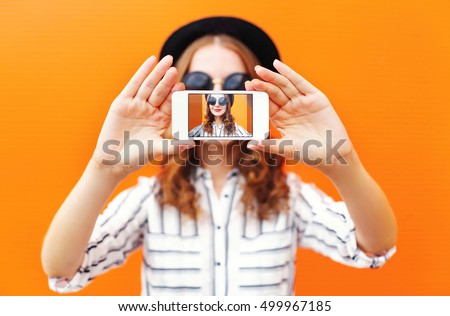 Fashion cool girl taking picture self portrait on smartphone over colorful orange background 