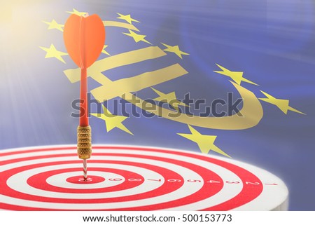 Closeup view of circular target marked with numbers and a red dart hits in the center, and an azure flag of Euro currency symbol with 12 yellow (gold) stars. An idea of popularity of Euro money.