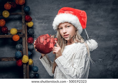 Cute blonde girl in Santa's hat holds red Christmas ball.