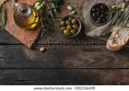 Green and black olives in tin cans with loaf of fresh bread and young olives branch on sackcloth, bottle of olive oil on clay board over old wood background. Overhead view with space for text