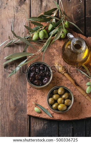 Green and black olives in tin cans, young olives branch and bottle of olive oil on clay board over old wood background. Overhead view with space for text