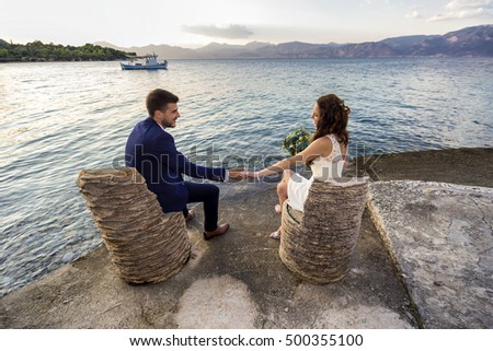 Young married couple sitting on chairs made of palm tree trunk, holding hands and looking at each other.