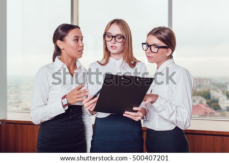 One Asian and two European business women standing against the window  study the documents