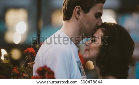 Attractive young man kisses woman's forehead while she leans to him between red flowers Royalty-Free Stock Photo #500478844