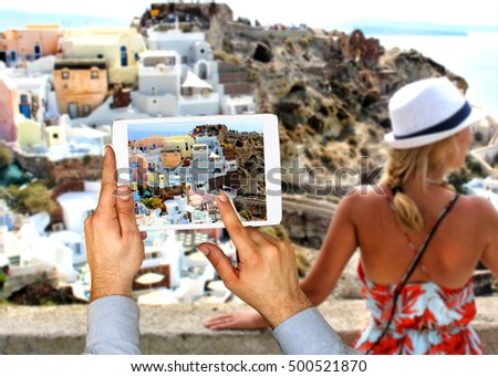 Tourist man taking picture with touch pad of woman on vacation relaxing in Fira town on Santorini island. Travel concept.  Europe summer travel destination in Greece, Caldera, Aegean sea.