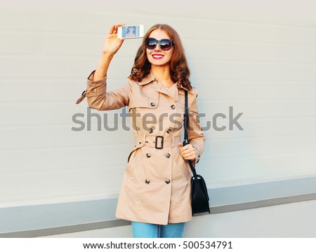 Fashion pretty smiling young woman taking picture self portrait on smartphone wearing a coat black handbag clutch in city over grey background