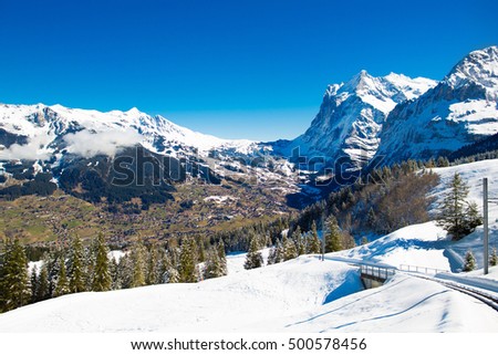 View from a train window in Switzerland on sunny spring day. Snow covered Alps mountain peaks. Railroad trip in Swiss Alps. Landscape with valley, village and mountains.