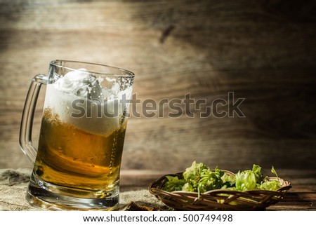 Big tankard of beer standing on empty wooden background with hops on table