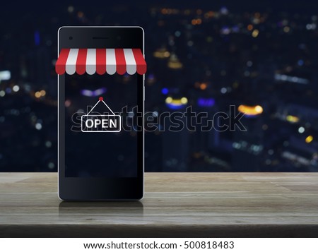 Modern smart mobile phone with on line shopping store graphic and open sign on wooden table over blur light city tower