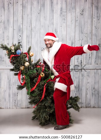 Picture of Bad Santa Clause riding on New Year Tree or Eve in studio. Happy man in Santa Clause clothes playing with New Year Eve.