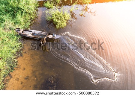 Asian fisherman on wooden boat casting a net for catching freshwater fish in nature river in the early morning before sunrise