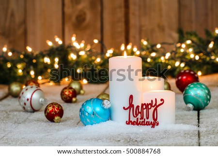 Christmas background with white candles, holiday ornaments, the words Happy Holidays and string of lights with green garland border in snow; red, teal blue, silver and gold rustic Christmas background
