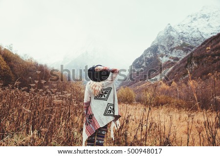 Boho woman wearing hat and poncho standing by the mountain. Cold weather, snow on hills. Winter hiking. Wanderlust. Royalty-Free Stock Photo #500948017