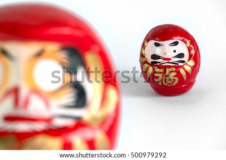 Closeup of a small red Daruma doll, with the typical missing eye, facing front, standing on the right of the picture, with a bigger one in the foreground, blurred by the DOF, on a white background.