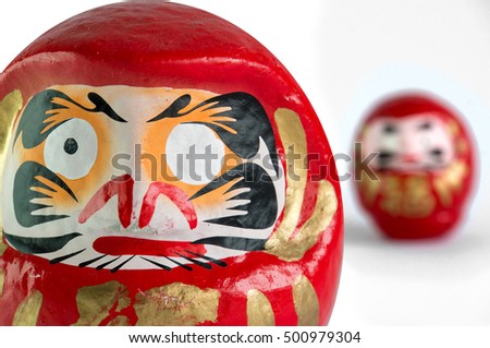 Closeup of a big red Daruma doll's head, with the typical missing eye, facing front, standing on the left of the picture, with a smallerone farther away, blurred by the DOF, on a white background.