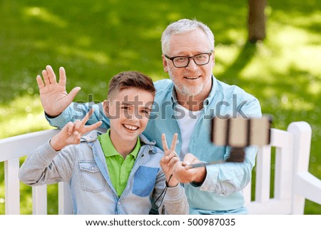 family, generation, technology and people concept - happy grandfather and grandson taking picture with smartphone selfie stick at summer park