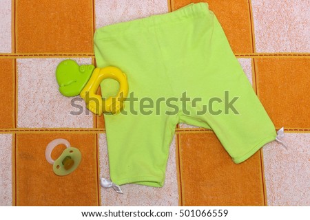 Green pants with white ribbons , green Hippo teether,  orthodontic nipple.Clothes for baby close up. Things for babies in orange and white plaid . Royalty-Free Stock Photo #501066559