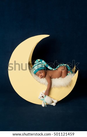 Studio portrait of a four week old newborn baby girl wearing a stocking cap and leg warmers. She is sleeping on a moon shaped posing prop and holding a crocheted Teddy bear.