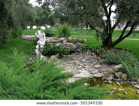 boy statue in antique style located near decorative stream on the background of decorative bushes and green grass