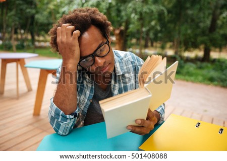 Thoughtful african young man sitting and reading book outdoors