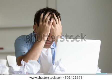 Portrait of a young man at the desk with a laptop, his hands closing his face. Business concept photo, lifestyle