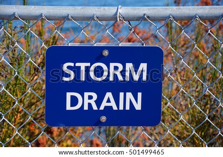 An image of a blue storm drain sign on a chain link fence.