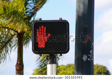 Illuminated Red Stop Don't Walk Hand Up Symbol Logo at Street Crosswalk with Palm Trees to the Left and Pole to the Right in the Afternoon