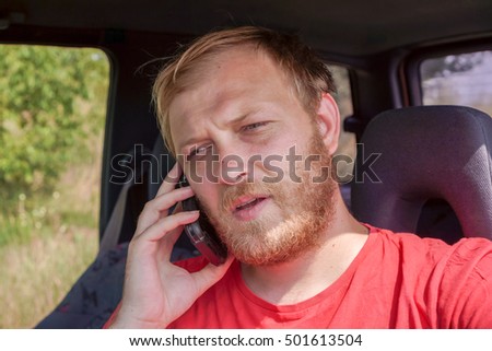 young blonde man speak telephone seating in his car, blonde man with blonde beard in red clothes speak with someone with telephone in right hand against car and green grass with shadows