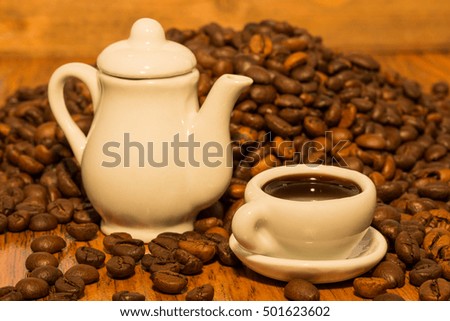 A tiny White teapot, a saucer and a cup of black coffee and background of coffee beans