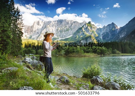 Beautiful young woman making sketches or writing in her notepad on the nature with Lago di Fusine lake with Mangart mountains in the background. Travel, Freedom, Lifestyle concept. Italy, Europe.