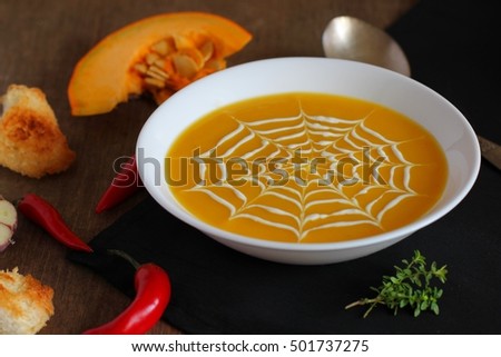 Fresh pumpkin soup with a picture of a spider web, cobweb  of cheese cream served on a board with garlic, pumpkin, chilli pepper and crouton on Halloween  .