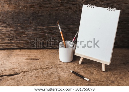 wooden easel with white paper, paintbrush and pencil on wooden background