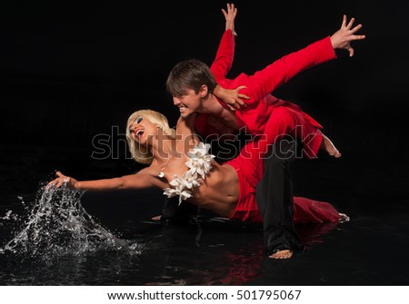  A pair of professional dancers in red dresses are dancing in the water. Emotional moment. Black background. Dance of love. Splashes of water. 