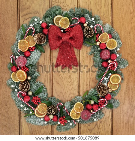 Christmas wreath with red bow decoration, dried fruit, candy canes, baubles, holly, mistletoe, pine cones and snow covered blue spruce fir over oak wood front door background.