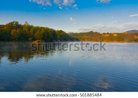Mountain lake for fishing against the background of green vegetation, blue morning sky in cirrus clouds and the rising sun. Beautiful reflection of objects in the water. Early morning. Western Ukraine