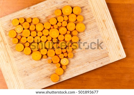 Chopped round and rectangular fresh orange colored carrots arranged in heart shape on wooden cutting board from top down perspective
