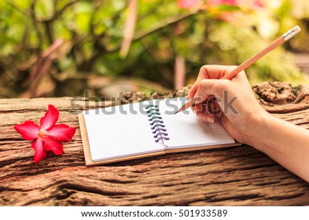 Hands of girl writing with a pencil in note book and Desert Rose standing on old wooden texture with green nature background. Closeup, Select focus. 