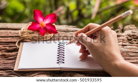 Hands of girl writing with a pencil in note book and Desert Rose standing on old wooden texture with green nature background. Closeup, Select focus. 