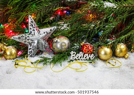 Christmas background with the word Believe, red and gold ornaments, silver star, colorful string of lights and Christmas tree garland border in snow; white copy space background