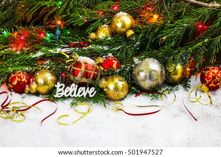 Christmas background with the word Believe, red and gold ornaments, colorful string of lights and Christmas tree garland border in snow; white copy space background
