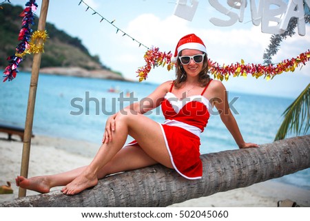 Cute blonde woman in red dress, sunglasses and santa hat sitting on palm tree at exotic tropical beach. Holiday concept for New Years Cards. Koh Samui. Thailand