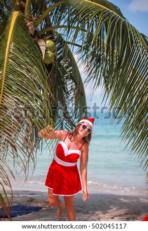 Cute blonde woman in red dress, sunglasses and santa hat stands at palm tree on exotic tropical beach. Holiday concept for New Years Cards. Koh Samui. Thailand