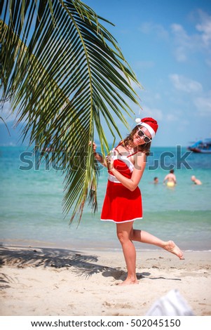 Cute blonde woman in red dress, sunglasses and santa hat stands at palm tree on exotic tropical beach. Holiday concept for New Years Cards. Koh Samui. Thailand