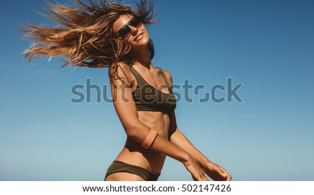 Portrait of beautiful young woman against blue sky with fluttering hair. Female model in bikini having fun outdoors.