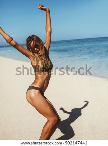 Full length portrait of happy young woman vacation having fun and dancing on the beach. Female model in bikini enjoying on the sea shore.