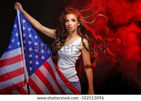 Young attractive girl holding US flag on the flagpole. She T-shirt and shorts with a print of the flag. Long curly hair flying. Portrait of a real patriot. Stars and Stripes. Smoke in the background.
