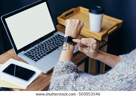 Woman using smart watch. Girl touching screen smart watch. Woman using digital gadget. In background, laptop with blank screen on wooden table, smartphone with black screen,cup of coffee, paper.