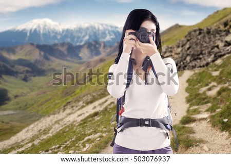 Young woman hiker taking pictures and looking at the camera with mountain view in the background